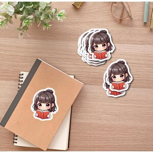 Puede incluir: Pegatinas que representan a una chica de dibujos animados con cabello casta&ntilde;o leyendo un libro rojo. Las pegatinas est&aacute;n sobre una superficie de madera, junto con un cuaderno y art&iacute;culos de papeler&iacute;a.