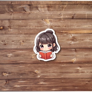 Puede incluir: Una pegatina con una ilustraci&oacute;n de estilo anime de una ni&ntilde;a con cabello oscuro en un mo&ntilde;o, leyendo un libro rojo. La pegatina tiene un borde blanco y est&aacute; sobre un fondo de madera.