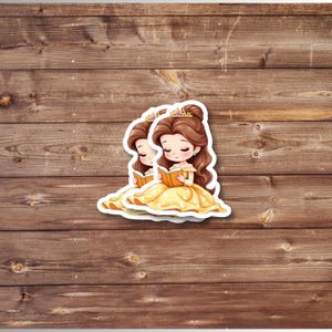 Puede incluir: Dos pegatinas con una ilustraci&oacute;n de dibujos animados de una princesa con un vestido amarillo, leyendo un libro. La princesa tiene el pelo casta&ntilde;o, una corona dorada y est&aacute; sentada. Las pegatinas est&aacute;n sobre un fondo de madera.