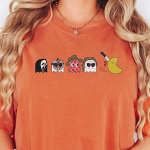 May include: Orange t-shirt featuring a row of cartoon ghosts dressed as horror movie characters, including Jason Voorhees and Freddy Krueger. A yellow Pac-Man ghost is being chased by a knife. The shirt has the text "YAM".