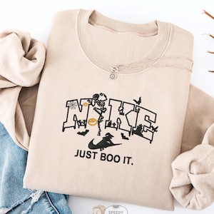 May include: Sand-colored sweatshirt with a black graphic that reads "NIKE" with Halloween-themed imagery, including a skeleton, bats, and pumpkins. Below the graphic, the text reads "JUST BOO IT." The sweatshirt is paired with blue jeans.