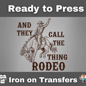 May include: A gray iron-on transfer with the text "Ready to Press" at the top. The design features a cowboy riding a horse with the words "AND THEY CALL THE THING RODEO" in brown. The bottom text reads "Iron on Transfers".