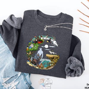 A gray sweatshirt with a colorful, illustrated design depicting various elements such as a castle, a dragon, and a unicorn, along with a necklace and a pair of jeans.