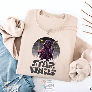 May include: A tan sweatshirt with a retro-style graphic of Darth Vader from Star Wars. The graphic features a cartoon version of Darth Vader holding a lightsaber with a TIE fighter flying overhead. The text "Star Wars" is printed below the graphic.