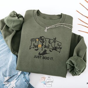 a green sweatshirt with a graphic design, a necklace, and some jeans, all laid out on a white background.