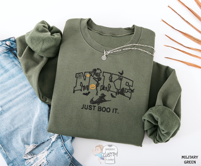 a green sweatshirt with a graphic design, a necklace, and some jeans, all laid out on a white background.