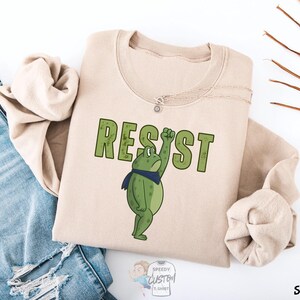 May include: Sand-colored sweatshirt with the word "RESIST" in green block letters. A cartoon frog wearing a blue scarf is raising its fist. The sweatshirt is folded next to a pair of blue jeans.
