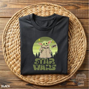 May include: Black t-shirt featuring a graphic of Grogu from Star Wars, set against a green and yellow circular background. The design includes the text "STAR WARS" in green. The shirt is folded on a woven mat.