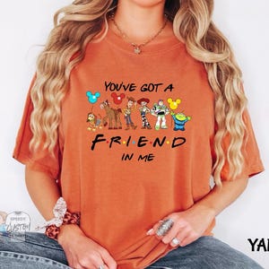 A person wearing an orange t-shirt with a graphic design featuring characters from the TV show "FRIEND IN ME" and the text "YOU'VE GOT A FRIEND IN ME".