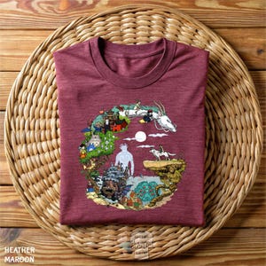 A maroon t-shirt with a colorful, whimsical illustration depicting various fantastical elements, including a dragon, castle, and people, all set against a serene landscape with a full moon.