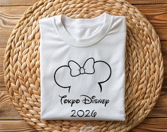 Puede incluir: Camiseta blanca con un diseño negro de orejas y lazo de Minnie Mouse, con el texto "Tokyo Disney 2026". La camiseta se muestra sobre una estera tejida sobre una superficie de madera. La palabra "WHITE" está impresa en la parte inferior.