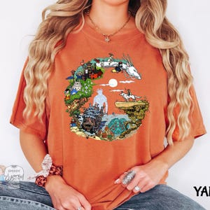 A woman wearing an orange t-shirt with a colorful graphic design depicting various elements such as a castle, a dragon, and a moon.