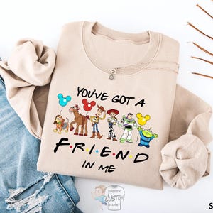 A beige sweatshirt with a graphic design featuring characters from the TV show "FRIENDS IN ME" and the text "YOU'VE GOT A FRIEND IN ME" printed on it.