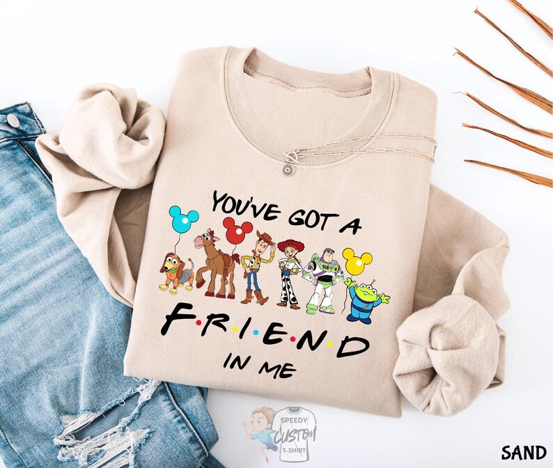 A beige sweatshirt with a graphic design featuring characters from the TV show "FRIENDS IN ME" and the text "YOU'VE GOT A FRIEND IN ME" printed on it.