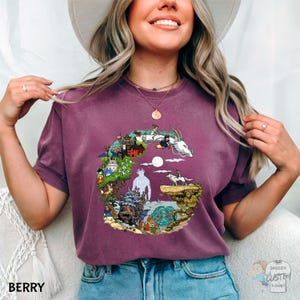 A smiling woman wearing a purple t-shirt with a colorful graphic design of a castle, a dragon, and a moon.