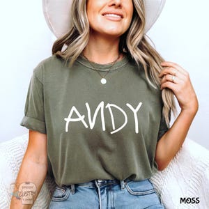 May include: Olive green t-shirt with the name "ANDY" in white script. The shirt is a relaxed fit and has rolled-up sleeves. The word "MOSS" is printed in the bottom right corner.