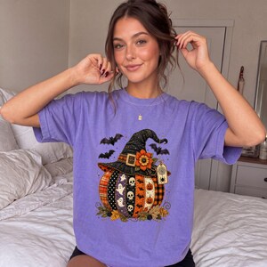 May include: A lavender short-sleeve t-shirt featuring a graphic print of a pumpkin wearing a witch hat. The pumpkin is decorated with vertical panels of skulls, ghosts, bats, and plaid patterns. A sunflower and a spider web tag adorn the hat.