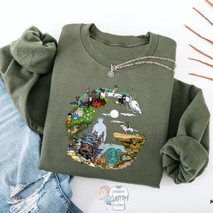 a green sweatshirt with a colorful, illustrated design on it, along with a pair of blue jeans and some leaves.