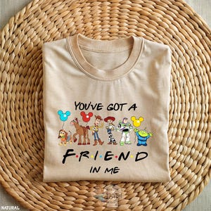 A beige t-shirt with a graphic design featuring characters from the animated film Toy Story, along with the text "You've got a FRIEND in ME" printed on it.