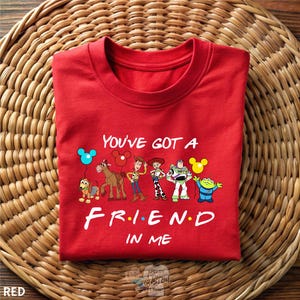 A red t-shirt with a cartoon character design and text printed on it, placed on a woven mat.