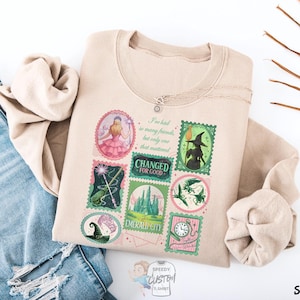 May include: Sand-colored sweatshirt featuring a collage of postage stamp-style graphics with images and text from the Wizard of Oz. The text includes "I've had so many friends but only one that mattered" and "Emerald City". A silver necklace is draped on the shirt.