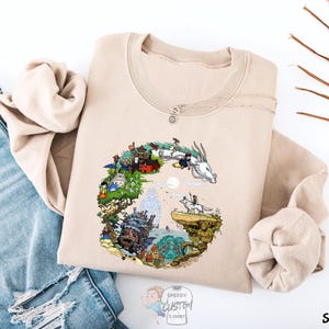 A beige sweatshirt with a colorful illustration of a dragon and various other elements, such as buildings, trees, and a castle, is displayed on a white background.