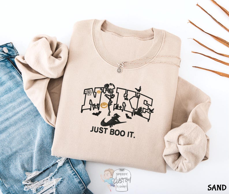 A beige sweatshirt with a graphic design and text printed on it, along with a pair of blue jeans and some dried leaves.