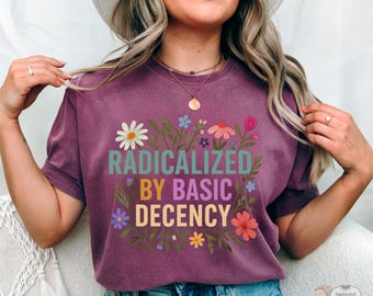May include: Berry-colored t-shirt with the text "RADICALIZED BY BASIC DECENCY" in a floral design. The shirt has a crew neck and short sleeves. The word "BERRY" is printed in the bottom left corner.