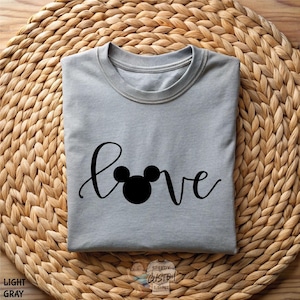 May include: Light gray t-shirt with the word "love" in a cursive font, with a Mickey Mouse head replacing the "o". The shirt is folded and rests on a woven, light brown surface. The words "Light Gray" are printed on the bottom left.