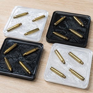 May include: Four square resin coasters featuring embedded brass bullet casings. Two coasters are clear with a white marbled effect, and two are solid black with a textured surface. Each coaster contains four metallic gold-toned casings.