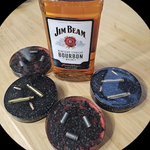 May include: A bottle of Jim Beam Kentucky Straight Bourbon Whiskey positioned behind three circular resin coasters. Each coaster features a dark, textured base embedded with metallic bullet casings and spent brass cartridges.