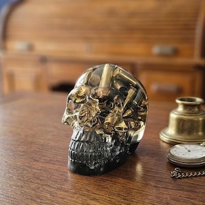 May include: A clear resin skull sculpture containing various metallic bullet casings embedded within the material. The skull features a smooth, polished surface with visible brass and copper-toned cartridges throughout the transparent structure.