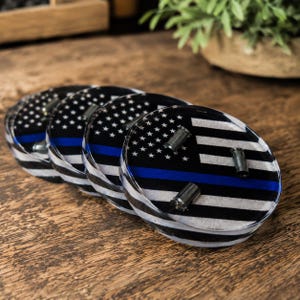 May include: A set of four circular coasters featuring a black and white American flag design with a horizontal blue stripe. Each coaster is encased in a clear, glossy material and includes three small, metallic bullet casing accents on the surface.