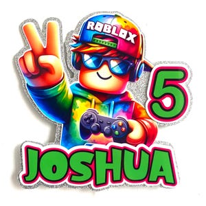 May include: A colourful cake topper featuring a Roblox character wearing a tie-dye shirt, sunglasses, and a hat that says "Roblox". The character is holding a video game controller and giving a peace sign. The topper also includes the number "5" and the name "Joshua" in green letters.