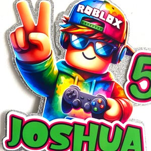 May include: A colourful cake topper featuring a Roblox character wearing a blue and red hat with the word "Roblox" on it. The character is holding a video game controller and has a green and pink name tag that says "Joshua".