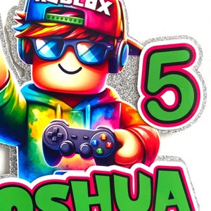 May include: A colourful Roblox character cake topper with a green and pink 5 and the name "Joshua" in green letters. The character is wearing a blue and pink hat with the word "Roblox" on it, sunglasses, and headphones. The character is holding a video game controller.