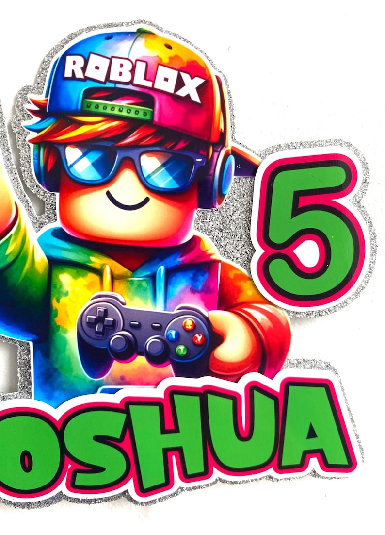 May include: A colourful Roblox character cake topper with a green and pink 5 and the name "Joshua" in green letters. The character is wearing a blue and pink hat with the word "Roblox" on it, sunglasses, and headphones. The character is holding a video game controller.