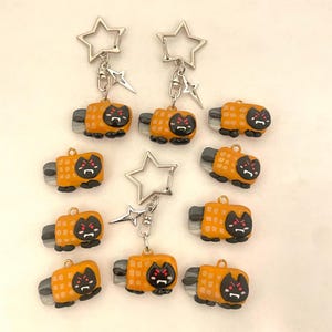 May include: Nine orange and black cat-shaped keychains with a star-shaped charm. Each keychain has a silver metal ring and a black cat face with red eyes.