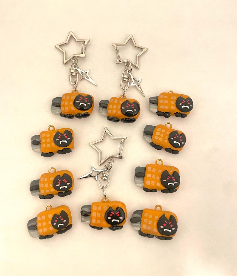May include: Nine orange and black cat-shaped keychains with a star-shaped charm. Each keychain has a silver metal ring and a black cat face with red eyes.