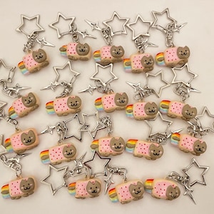 May include: A collection of 20 keychains featuring a pink and white cat with a rainbow on its back. The cat is holding a slice of bread with a pink frosting and sprinkles. Each keychain is attached to a silver star-shaped charm and a silver chain.