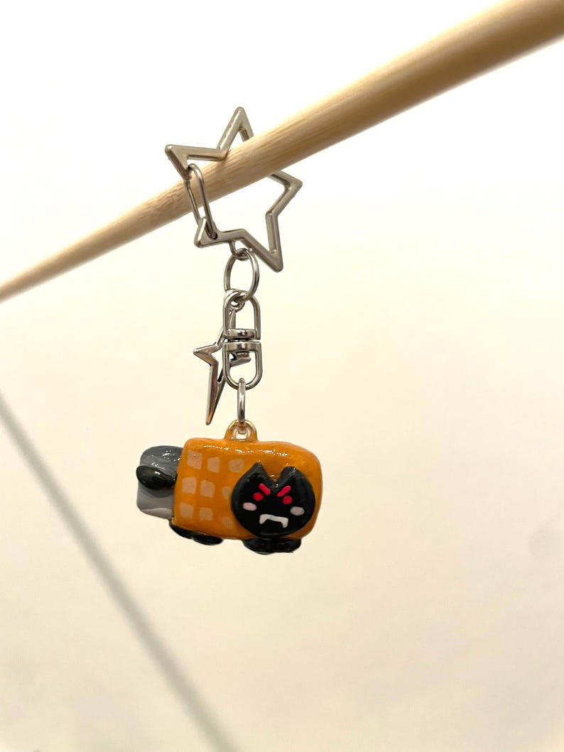 May include: A yellow and black cat-shaped keychain with a waffle pattern on its back. The keychain is attached to a silver star-shaped charm and a silver chain.