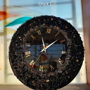 Può includere: Un orologio da parete rotondo nero con numeri romani e lancette dorate. Il quadrante dell'orologio è nero e il bordo esterno è decorato con pietre nere e dettagli dorati. Il testo "Clock 1" è in alto.