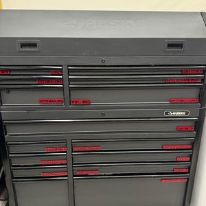 May include: A black Husky rolling tool chest with multiple drawers labeled for various tools, including wrenches, screwdrivers, and sockets. The chest has a top compartment and is on wheels for easy mobility.