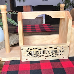 Puede incluir: Un organizador de cocina de madera con un portarrollos de papel, un soporte para utensilios y un estante. El organizador tiene las palabras "Grillin, Chillin, Refillin" en una fuente decorativa. Se encuentra sobre un mantel individual a cuadros rojos y negros.