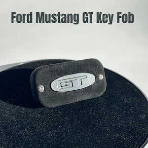 May include: A black Ford Mustang GT key fob with a silver GT emblem. The key fob is rectangular with rounded edges and is made of a soft, suede-like material. The text "Ford Mustang GT Key Fob" is displayed above the key fob.