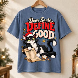 May include: A blue short-sleeve t-shirt featuring a graphic of a black and white cat wearing a red Santa hat. The cat holds a scroll labeled NAUGHTY LIST. Text above reads Dear Santa, DEFINE GOOD. Yellow stars and a cookie are also visible.