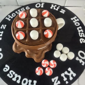May include: A chocolate-colored waffle-shaped mug with white marshmallows and red and white striped candies. The mug sits on a black circular base with the text "House Of Kiz" in white.