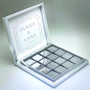 May include: A silver gift box with a hinged lid, filled with square chocolates. The box lid features the names "JAMES & ANNA" and the date "10-12-2035" inside a diamond-shaped design. Each chocolate square is individually wrapped.