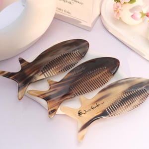 May include: Three fish-shaped combs with a natural, marbled pattern in shades of brown and tan. One comb has the name "Linda" engraved, and another has a floral design with the name "Anuka". The combs are displayed on a white surface.