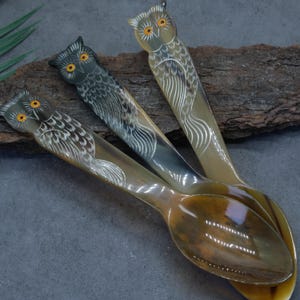 May include: Three spoons crafted from polished horn, each featuring a carved owl handle with etched feather details and painted yellow eyes. The spoons exhibit natural color variations ranging from dark charcoal to light amber tones.